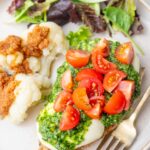 Chicken breast topped with mozarella, pesto, and tomatoes on a beige plate with cauliflower and a salad.