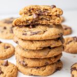 A stack of golden-brown Biscoff chocolate chip cookies, with the top cookie broken in half to show gooey chocolate inside. Other cookies are scattered in the background.