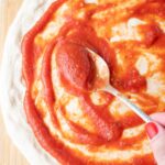 A hand uses a spoon to spread pizza sauce over a round pizza dough on a floured wooden surface, with fresh basil leaves in the top corner.